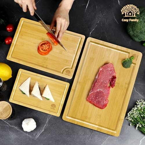 Hands slicing tomato on bamboo cutting board set with steak and sandwiches.