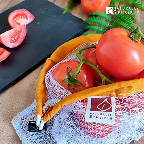 Fresh tomatoes in a mesh bag on a table with sliced tomatoes on a slate board.